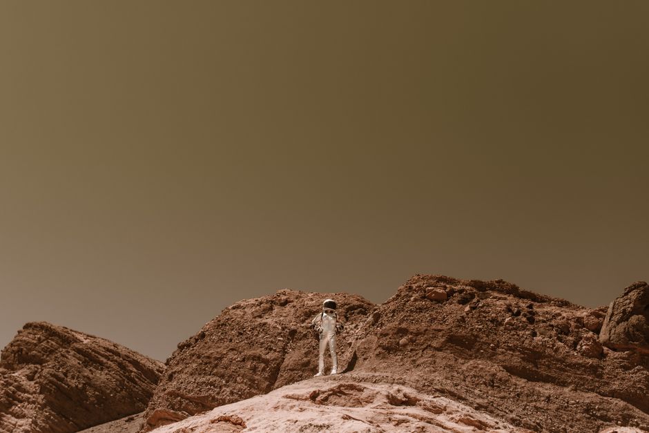 Astronaut in a silver space suit standing on barren rocky terrain, resembling Mars.